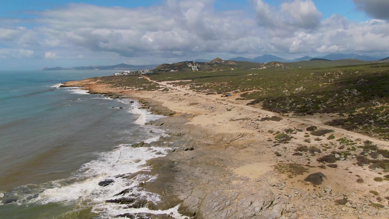 Aerial view over Buzzards Beach East Cape BCS Mexico with ocean surf at left, sandy rocky shoreline below, green hills beyond, cloudy sky, and no visible people.