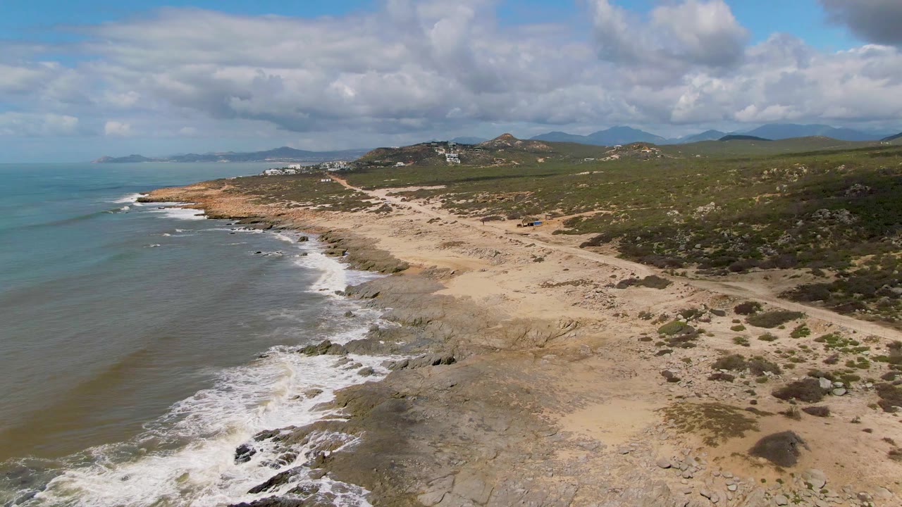 Drone view along Buzzards Beach East Cape BCS Mexico showing foamy waves on the left, pale sand and rock shelves below, inland dirt tracks, green hills, and no visible people.