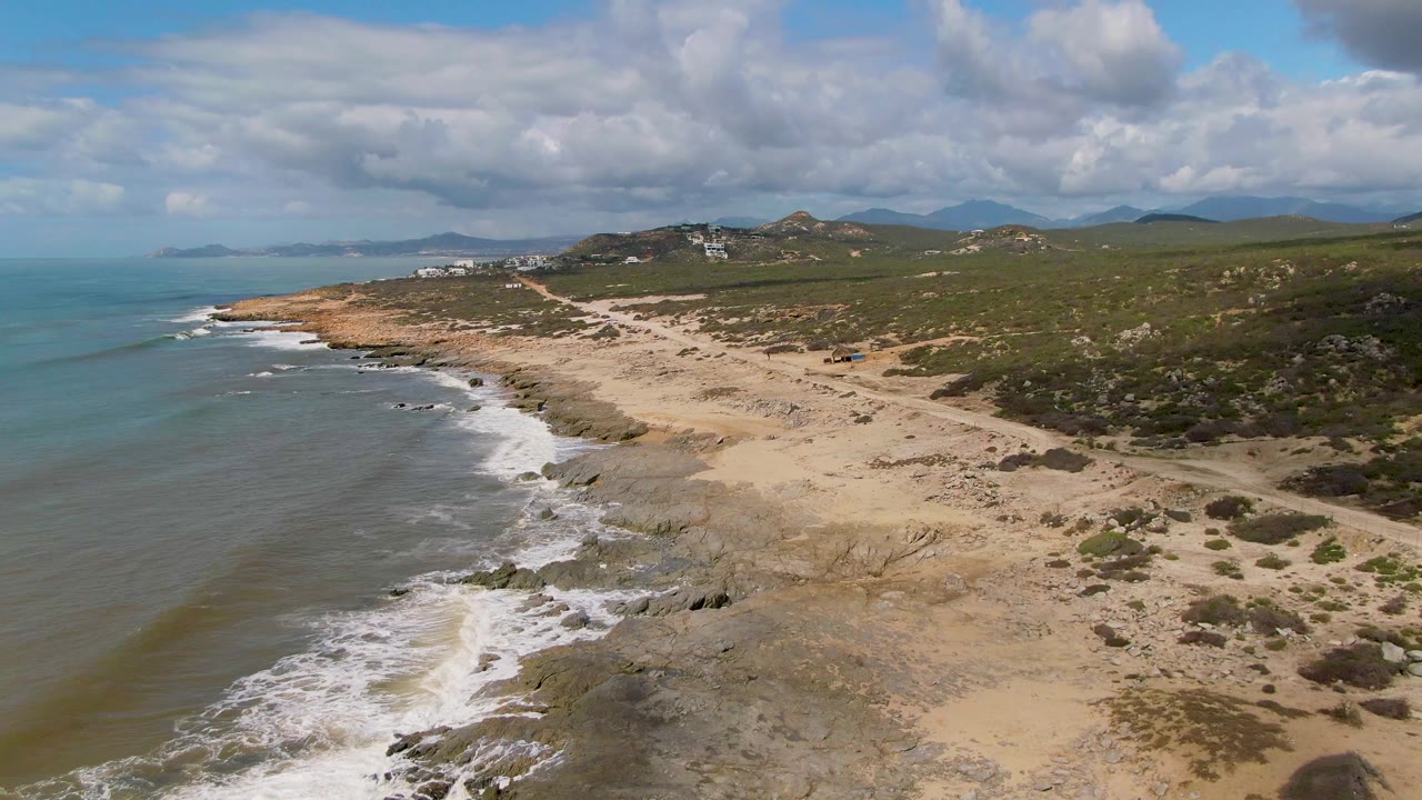 Oblique aerial frame at Buzzards Beach East Cape BCS Mexico with the surf line curling along the left, tan sand and gray rock in the foreground, green hills behind, and no visible people.