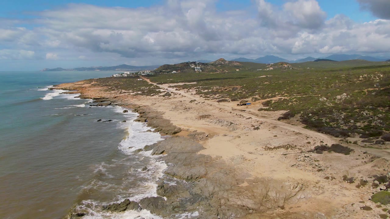 Opening aerial frame over Buzzards Beach East Cape BCS Mexico with surf on the left, tan rocky shoreline below, green scrub hills beyond, distant buildings, and no visible people.