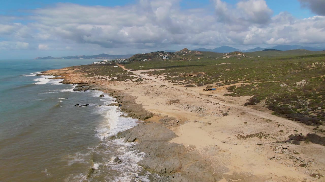 Aerial frame over Buzzards Beach East Cape BCS Mexico with waves breaking across the left shoreline, pale sand and exposed rock in the center, inland scrub and hills in the distance, and no visible people.