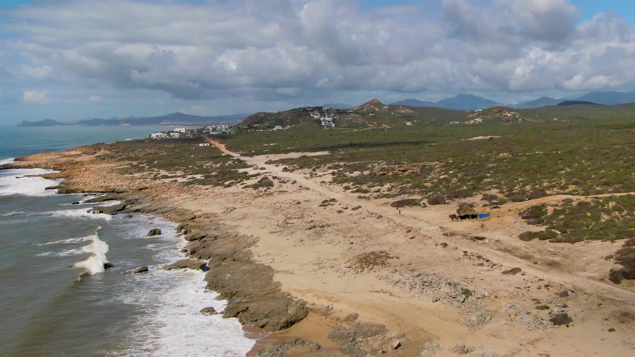 Opening aerial view of Buzzards Beach in Mexico with surf along the left edge, a sandy rocky shore, low green brush, distant hills, cloud cover, and no visible people.