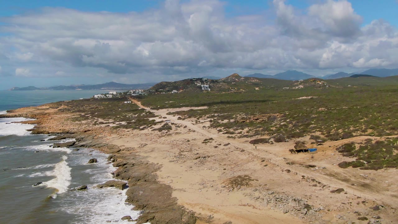 Surf breaks beside a sandy beach with a dirt road and hillside buildings inland.
