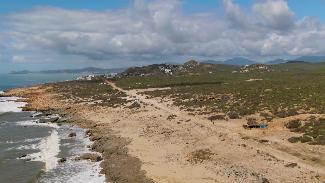 Mid-clip aerial view of Buzzards Beach in Mexico with the ocean on the left, waves curling onto rocks, sandy flats across the center, scrub-covered hills, distant buildings, and no visible people.