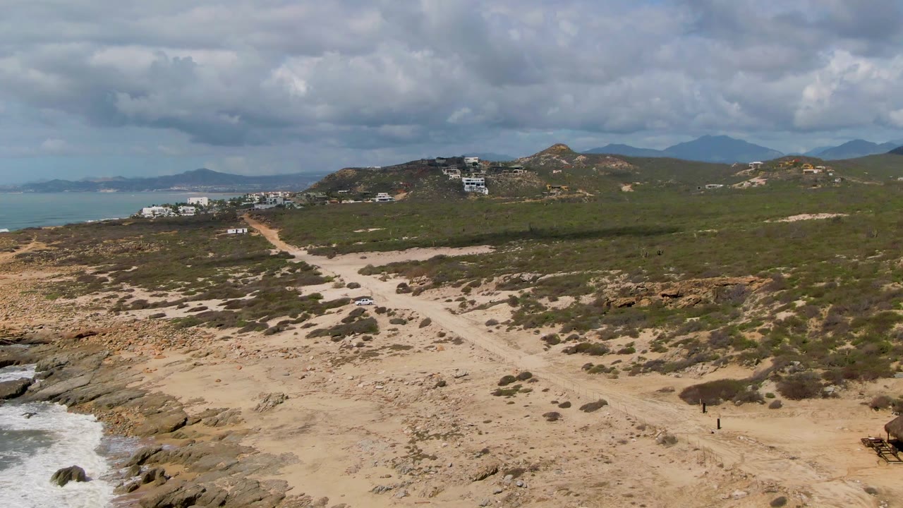 Opening frame of Buzzards Beach East Cape BCS Mexico showing a sandy dirt road through scrub and rock, ocean on the left, hillside buildings in the distance, and no visible people.