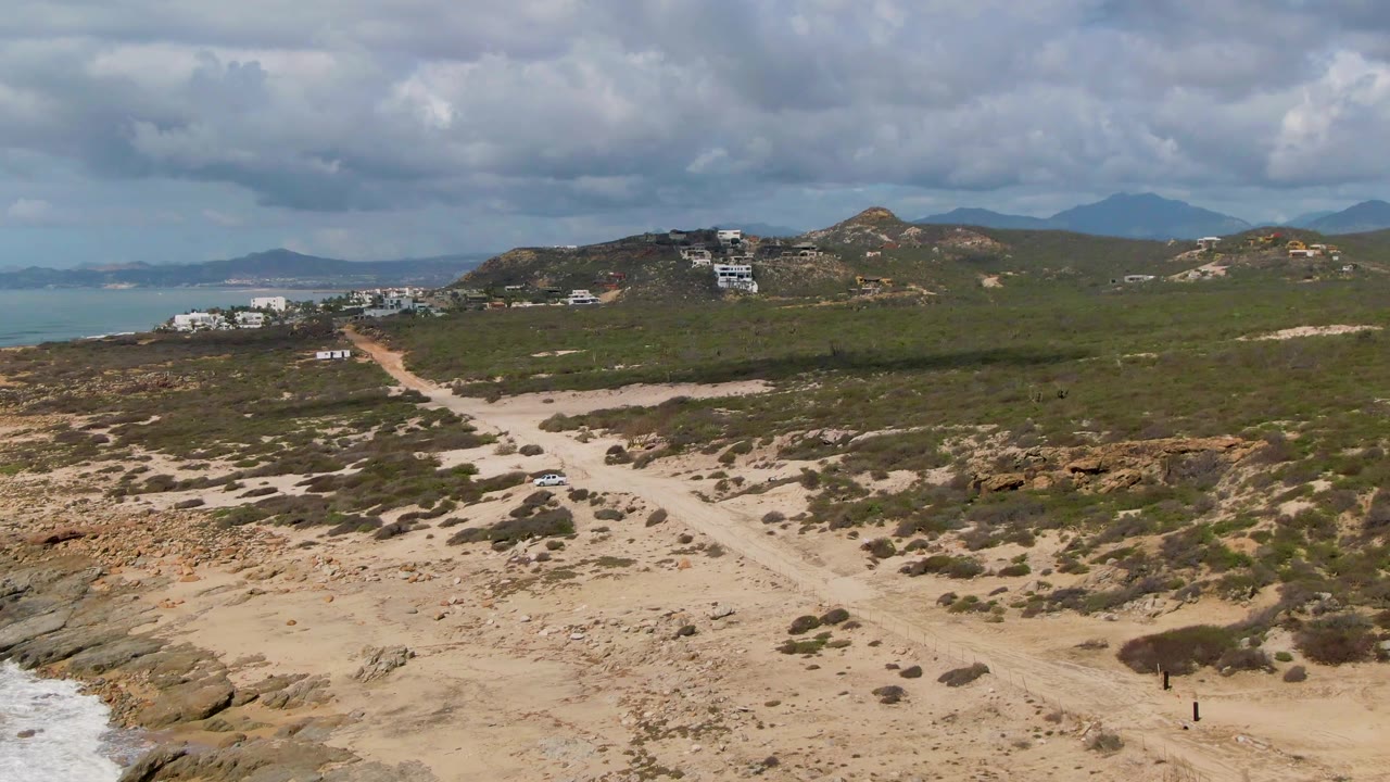 Sandy road crossing coastal scrub with hillside buildings beyond