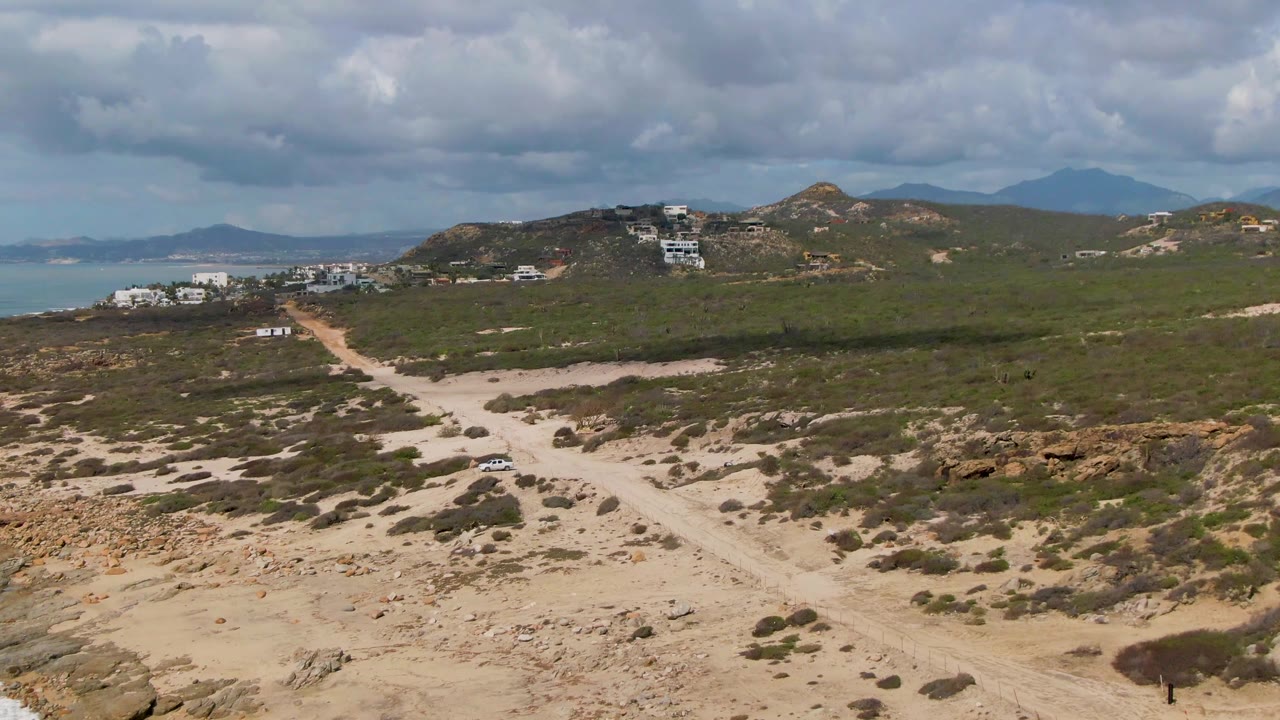 Wide aerial frame at Buzzards Beach East Cape BCS Mexico with rocky foreground, a pale dirt road leading inland, ocean visible at left, hills and buildings beyond, and no visible people.
