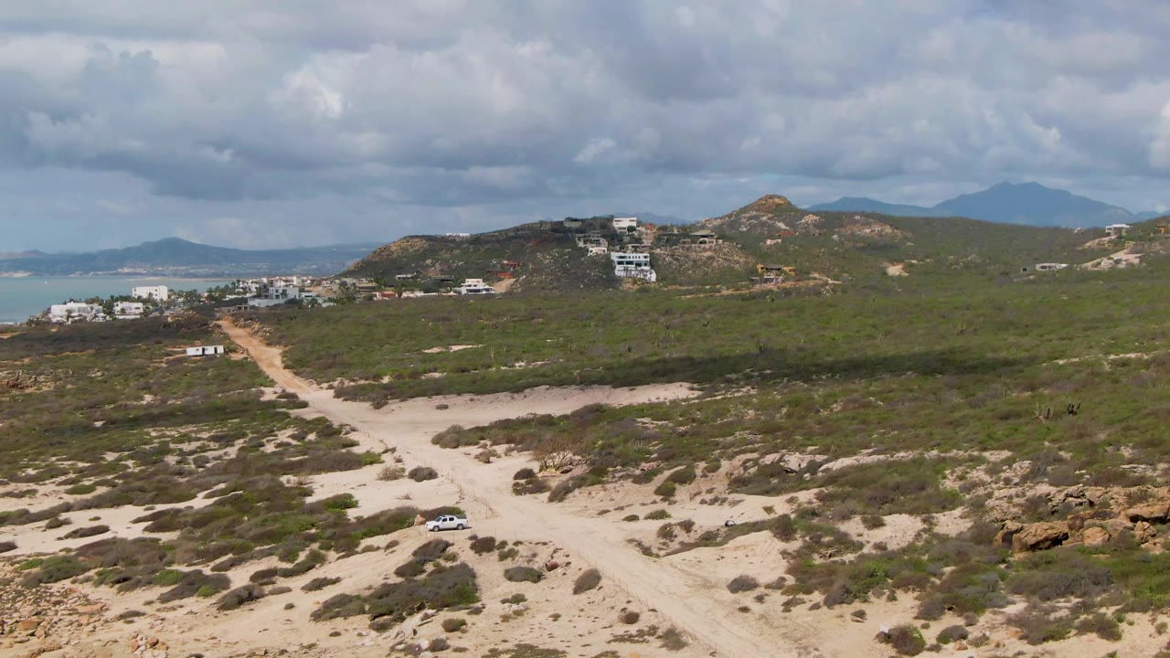 Aerial view at Buzzards Beach East Cape BCS Mexico showing a small car on a pale dirt road, green scrub, rocky hills, scattered buildings, distant sea, and no visible people.