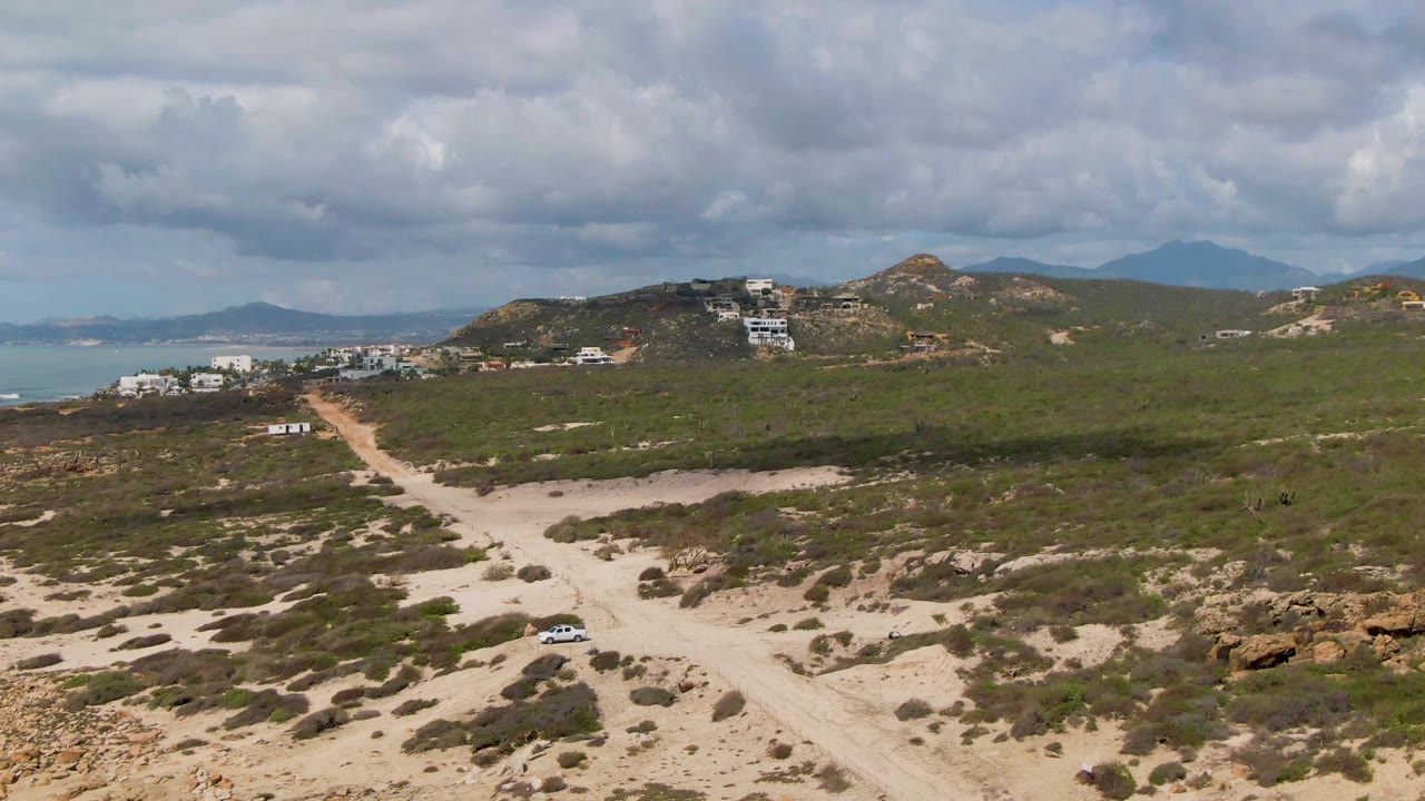 Aerial frame at Buzzards Beach East Cape BCS Mexico with the dirt road cutting diagonally through scrubland, a small car near the lower road, hillside buildings, distant water, and no visible people.