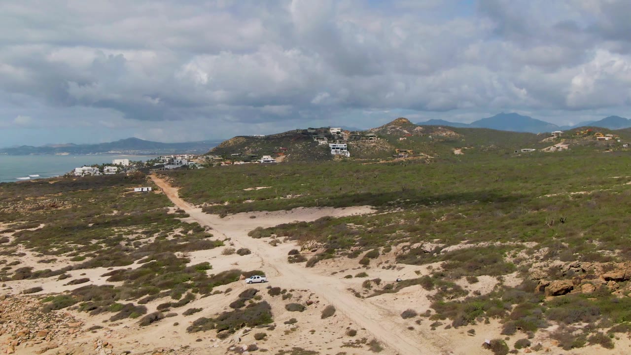 Aerial view at Buzzards Beach East Cape BCS Mexico showing the car on the lower winding road, broad green vegetation across the hills, distant ocean at left, layered clouds, and no visible people.