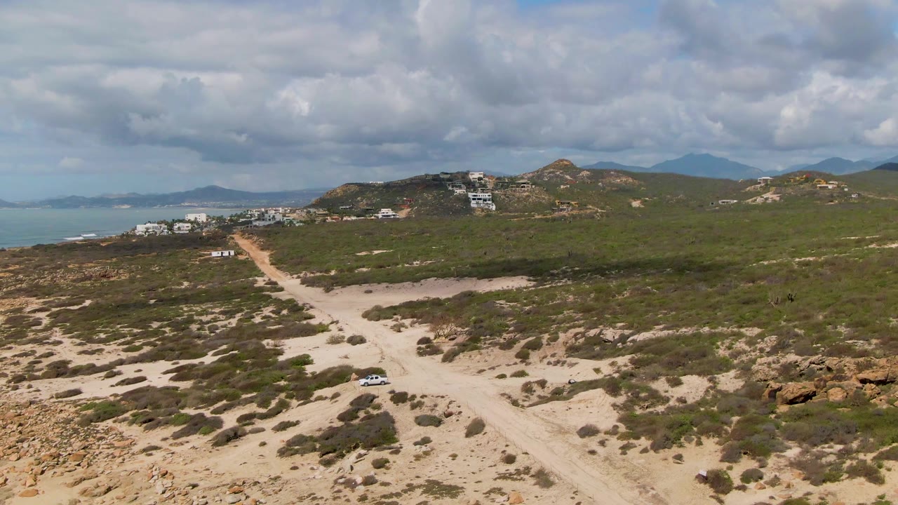 Winding dirt road through scrub with hills, buildings, and distant coast.