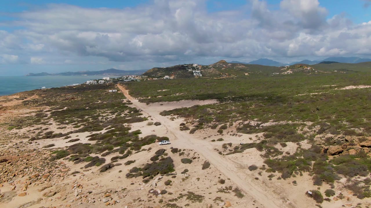 Aerial view at Buzzards Beach East Cape BCS Mexico showing a white vehicle on a sandy road through low shrubs, with ocean on the left, hills in the distance, and no visible people.