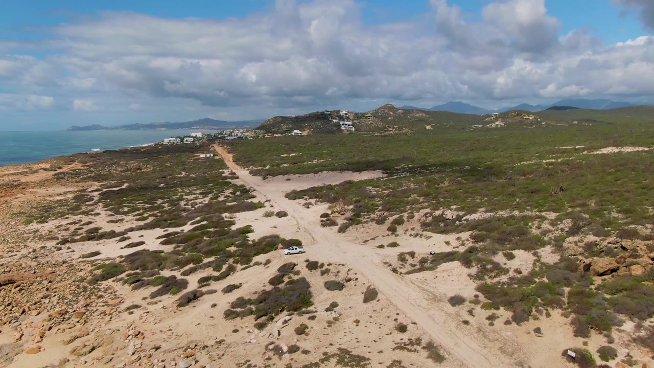 Aerial frame from Buzzards Beach East Cape BCS Mexico with the white vehicle continuing along the pale road, surrounded by green scrub, rocky ground, distant buildings, ocean, and no visible people.