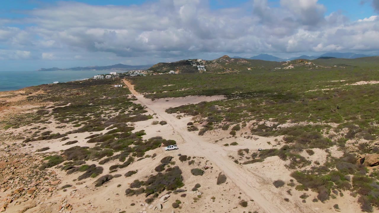 Buzzards Beach East Cape BCS Mexico aerial view showing the white vehicle lower in frame on the sandy road, with scrubland, rocky patches, ocean water to the left, hills, clouds, and no visible people.
