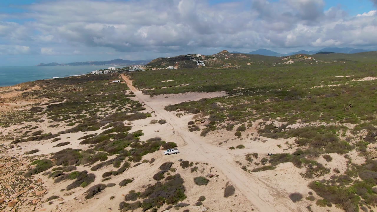 Wide aerial of a sandy road through coastal shrubs with ocean and hills.