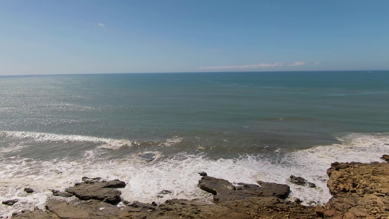 Ocean waves breaking over dark rocks below a blue horizon.