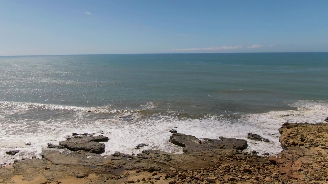 At Buzzards Beach in East Cape BCS, Mexico, a brighter band of whitewater breaks around uneven shoreline rocks while the ocean remains smooth beyond, with no visible people.