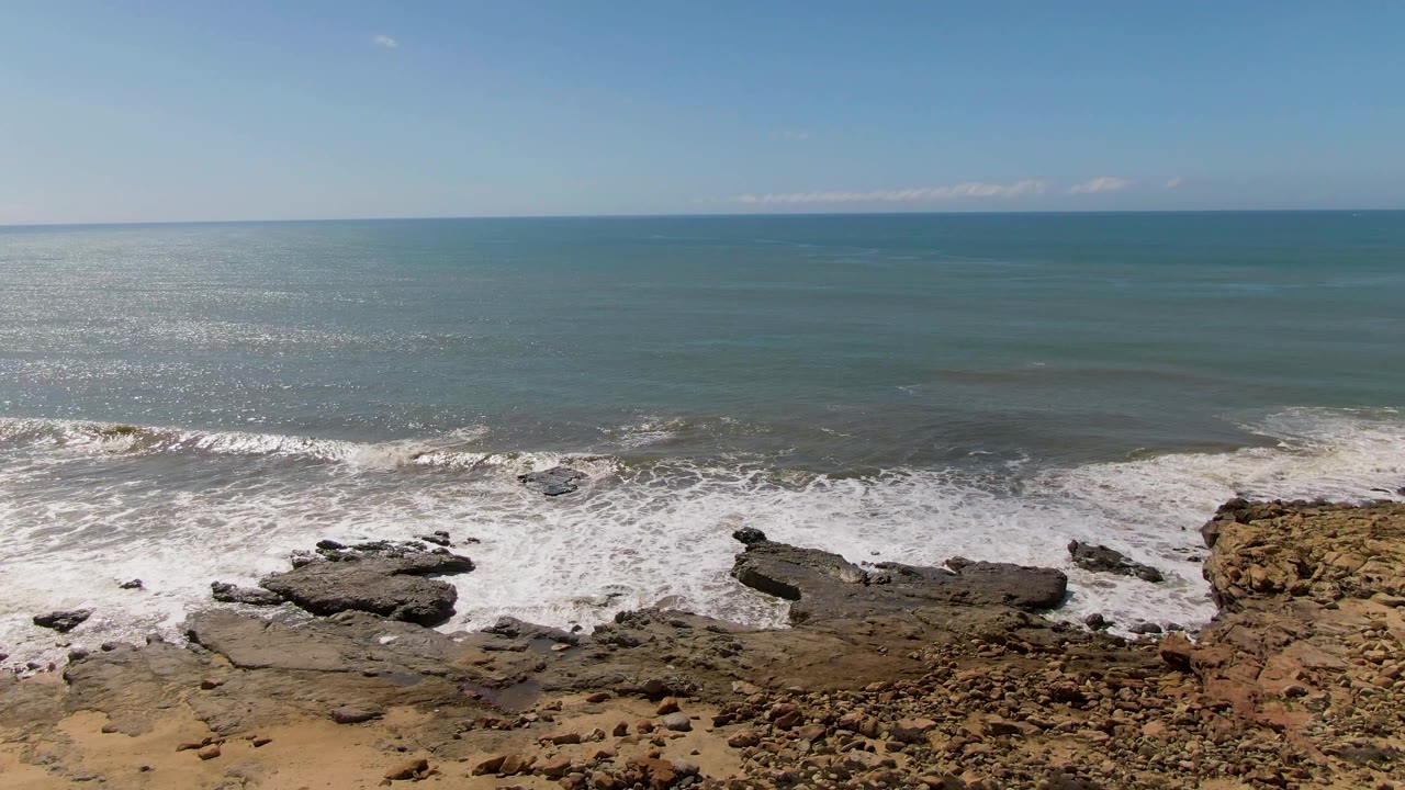 At 0:03, the foamy water pulls across the lower rocks at Buzzards Beach, leaving open blue-green ocean and a clean horizon above, with no visible people.