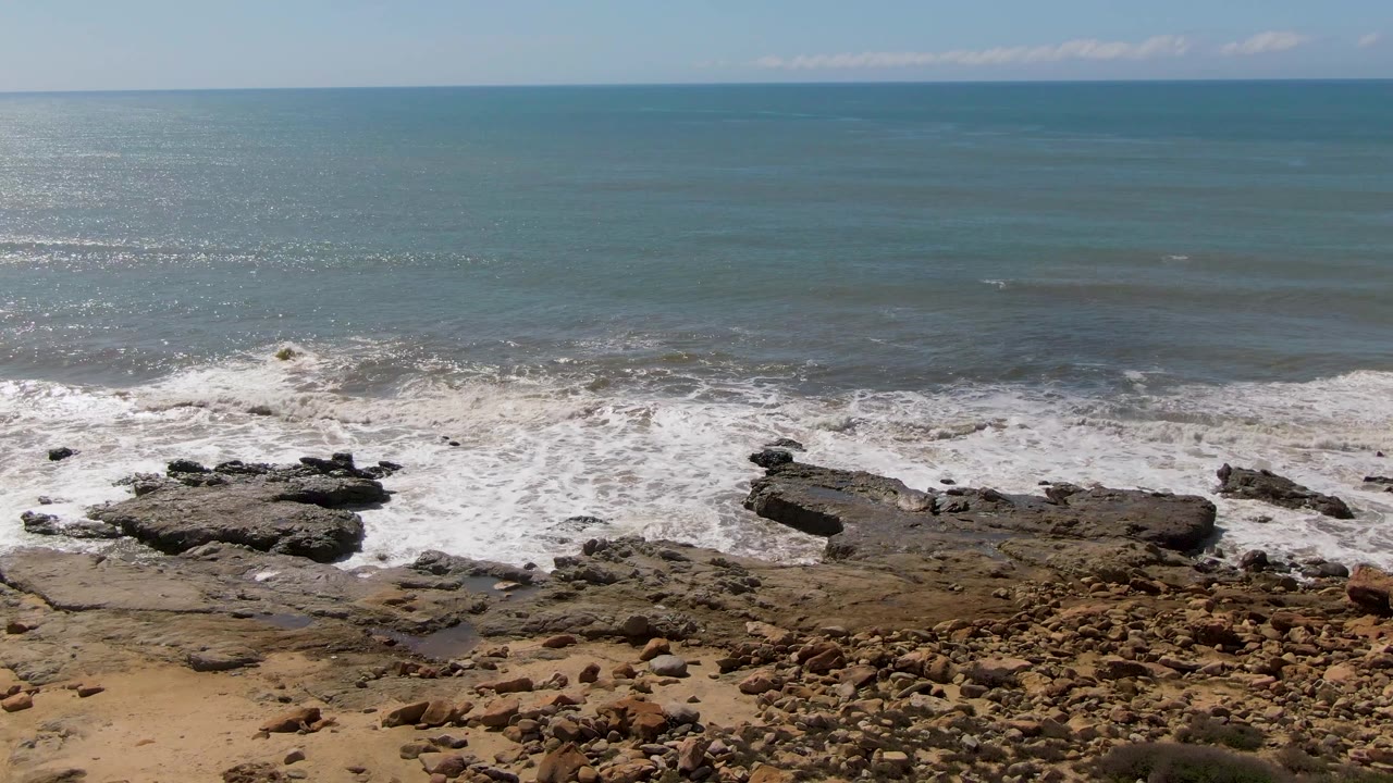 At Buzzards Beach East Cape BCS Mexico, a brighter band of foam pushes across flat shoreline rocks while the open blue-green sea stretches to the horizon, with no visible people.