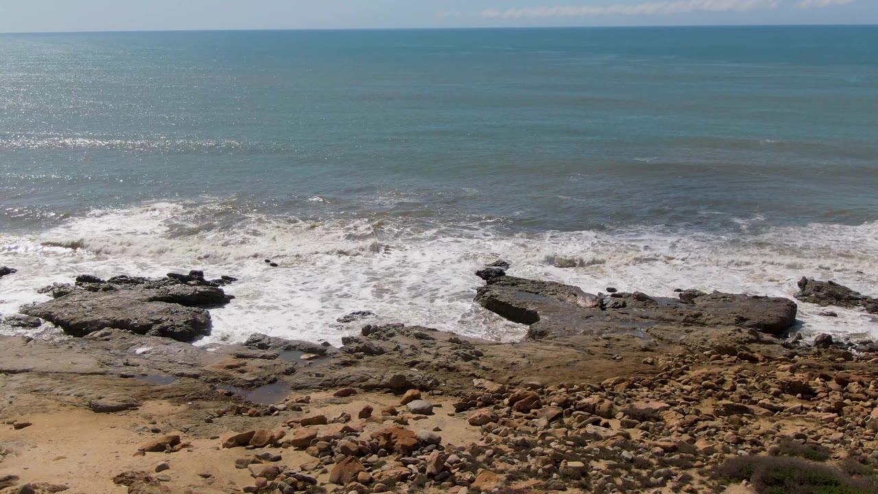 At Buzzards Beach East Cape BCS Mexico, foamy surf wraps around dark rock outcrops along a stone-strewn sandy edge beneath a wide blue ocean, with no visible people.