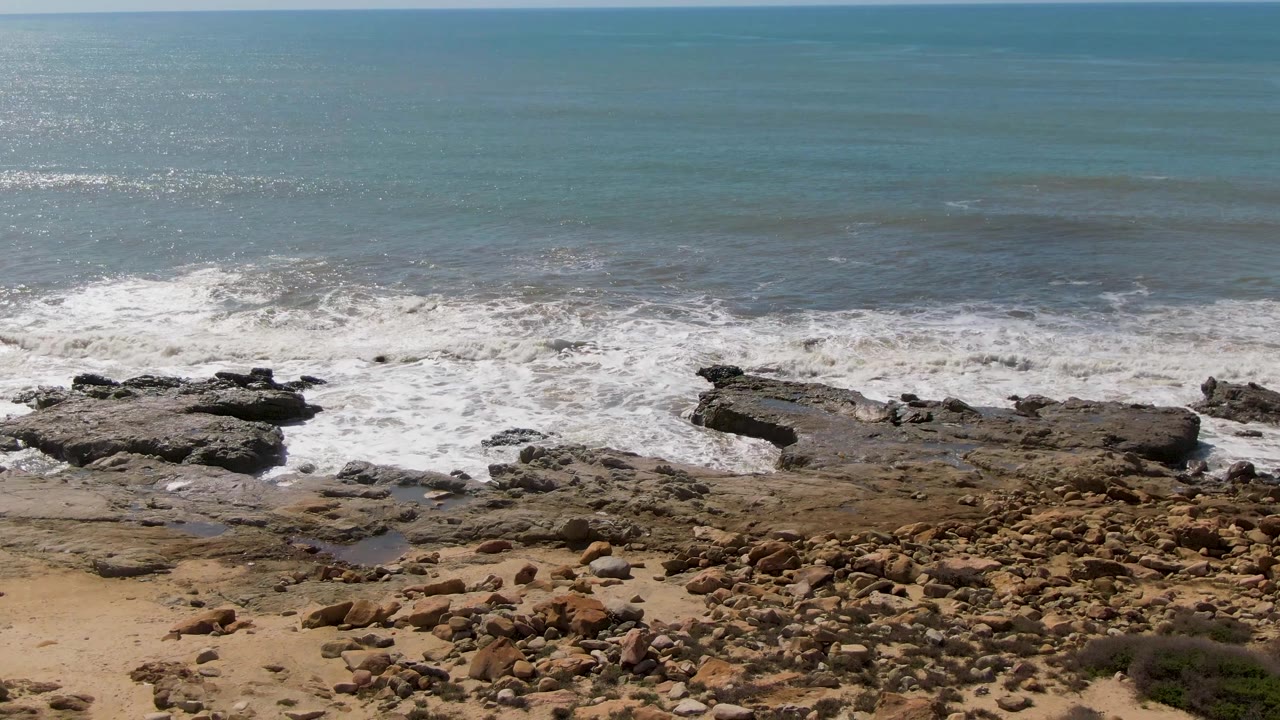 At Buzzards Beach East Cape BCS Mexico, the lower shoreline shows tan sand, rounded brown rocks, and white surf sliding across the rock shelf below the blue-green ocean, with no visible people.