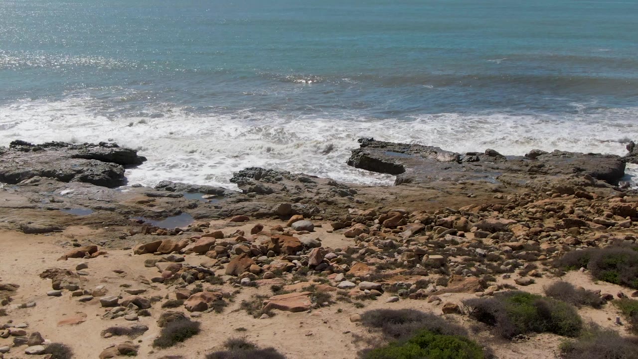 Aerial view of Buzzards Beach East Cape BCS Mexico with blue water, white waves breaking over dark rocks, tan sand, scattered stones, low shrubs, and no visible people.