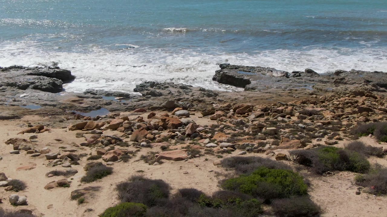 Aerial shoreline frame from Buzzards Beach East Cape BCS Mexico showing turquoise-blue water, white surf, flat dark rocks, tan beach stones, scrub vegetation, and no visible people.
