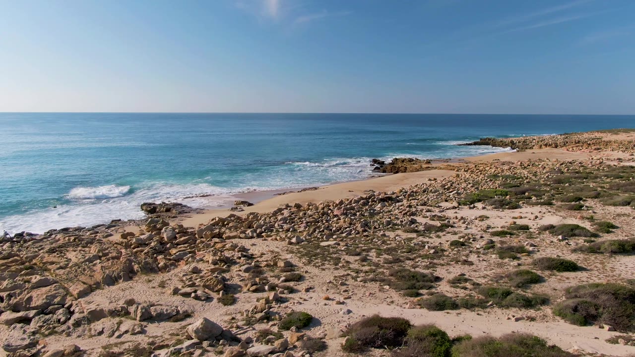 Fourth frame at Buzzards Beach East Cape BCS Mexico with the surf lower in frame, rocky shoreline across the middle, sparse green shrubs on sand, and no visible people.
