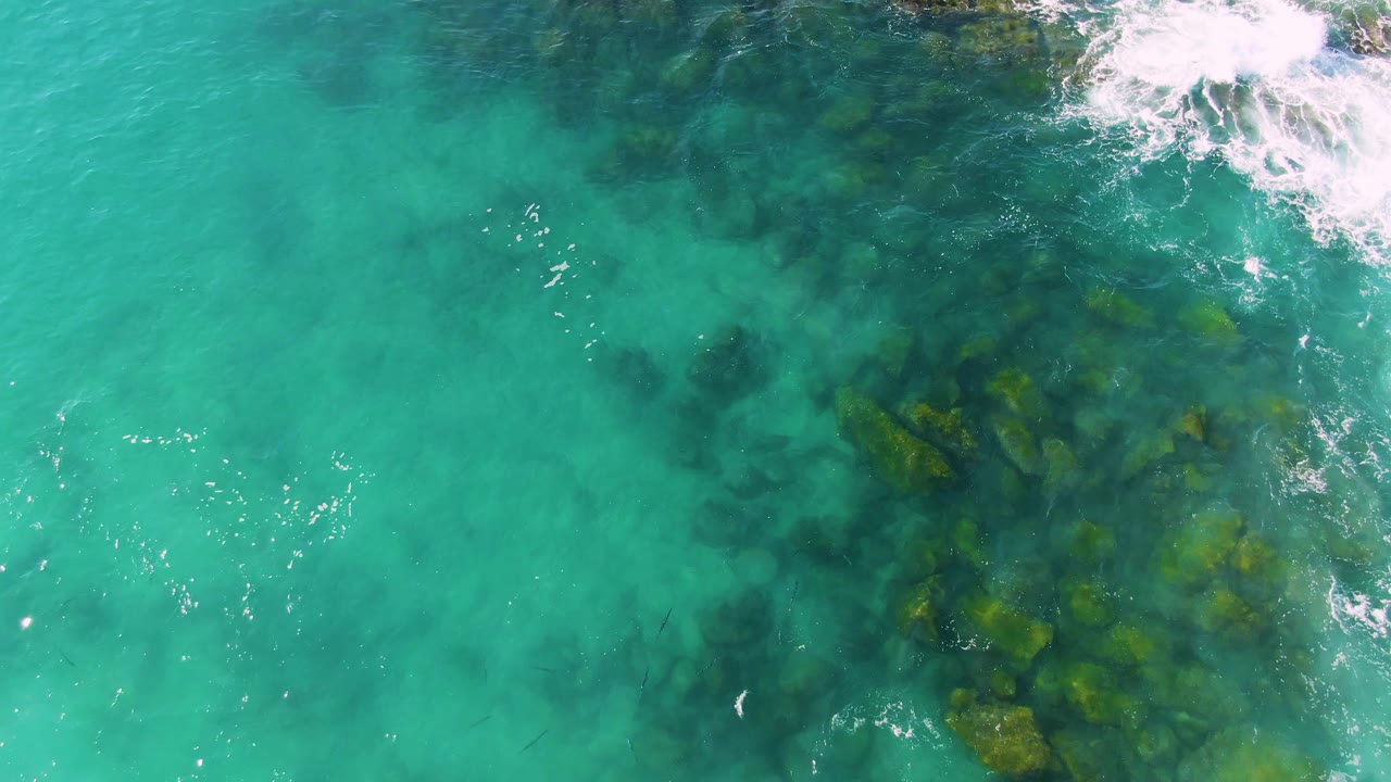 Turquoise ocean water over dark underwater rocks with white foam near the upper right.