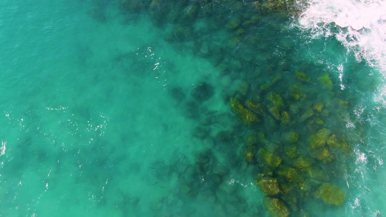 Aerial frame of green-blue water at Buzzards Beach East Cape BCS Mexico, with a cluster of submerged rocks shifted toward the right side and no visible people.