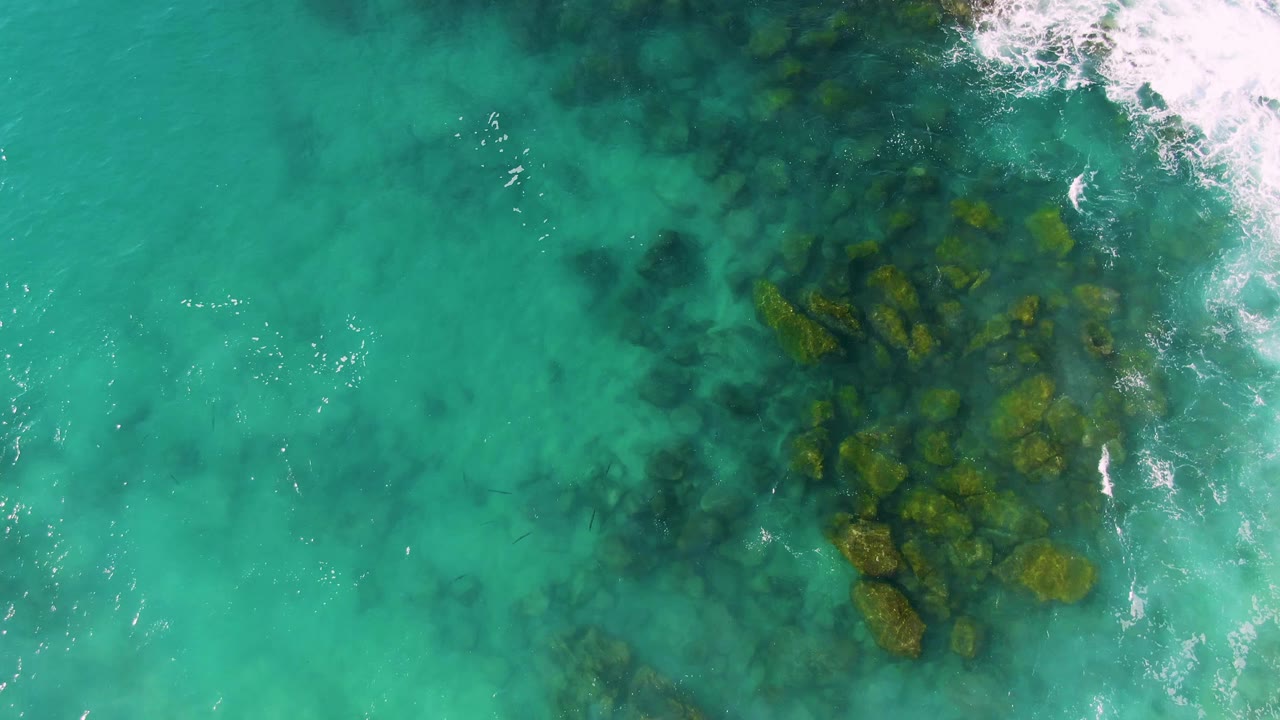 Top-down drone view over Buzzards Beach East Cape BCS Mexico, showing a denser rocky patch beneath turquoise water, foam touching the upper right border, and no visible people.