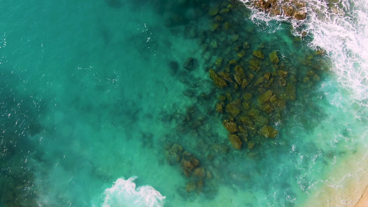Overhead view at Buzzards Beach East Cape BCS Mexico showing turquoise water, dark submerged rocks, white surf at the upper right, and no visible people.