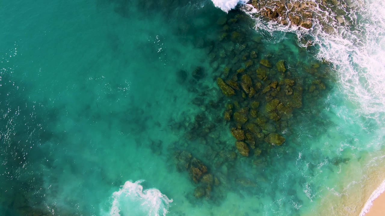 Aerial frame at Buzzards Beach East Cape BCS Mexico with submerged rock clusters centered under clear turquoise water and white foam entering from the upper right, with no visible people.