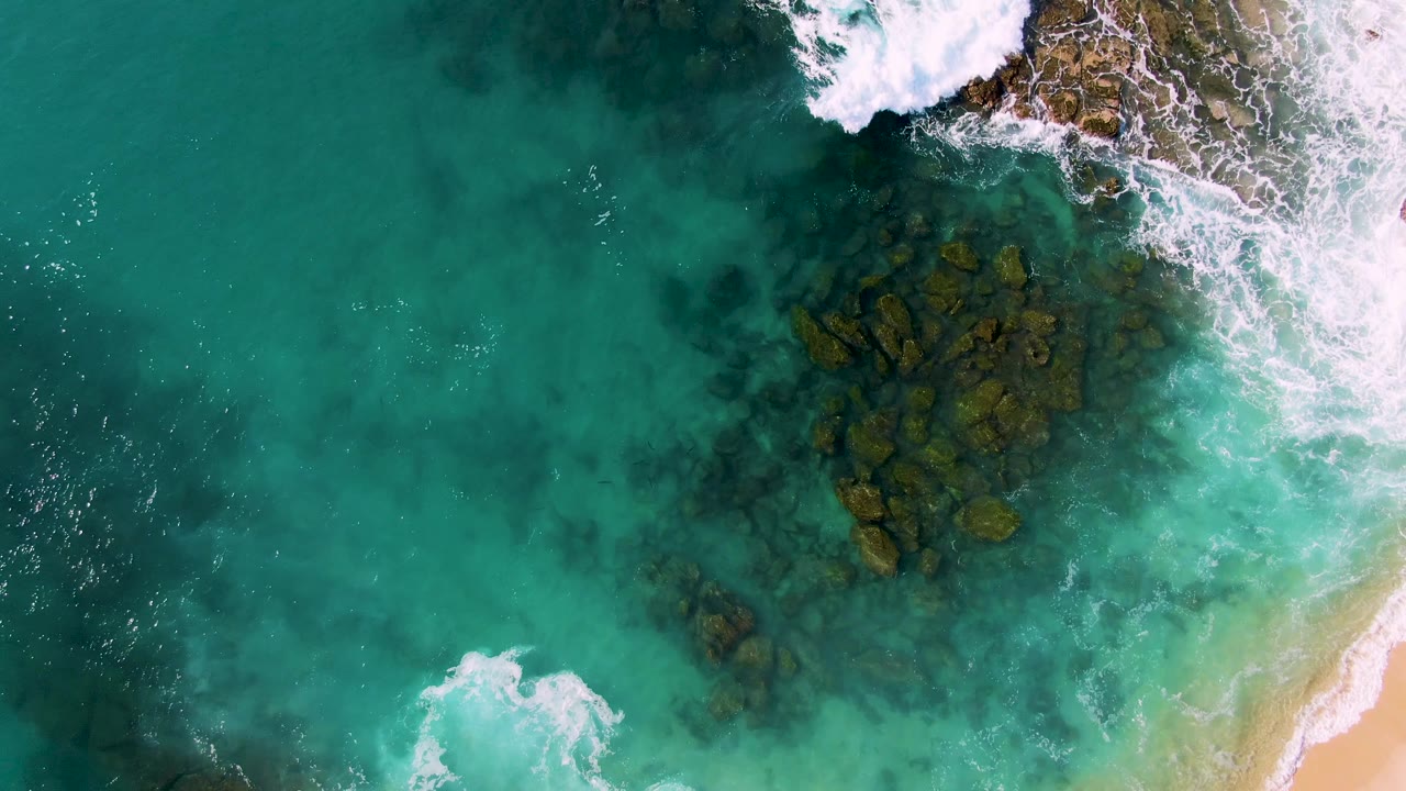 Drone view at Buzzards Beach East Cape BCS Mexico showing dark rocks under green-blue water, surf breaking at the top right, a narrow sandy beach edge at lower right, and no visible people.