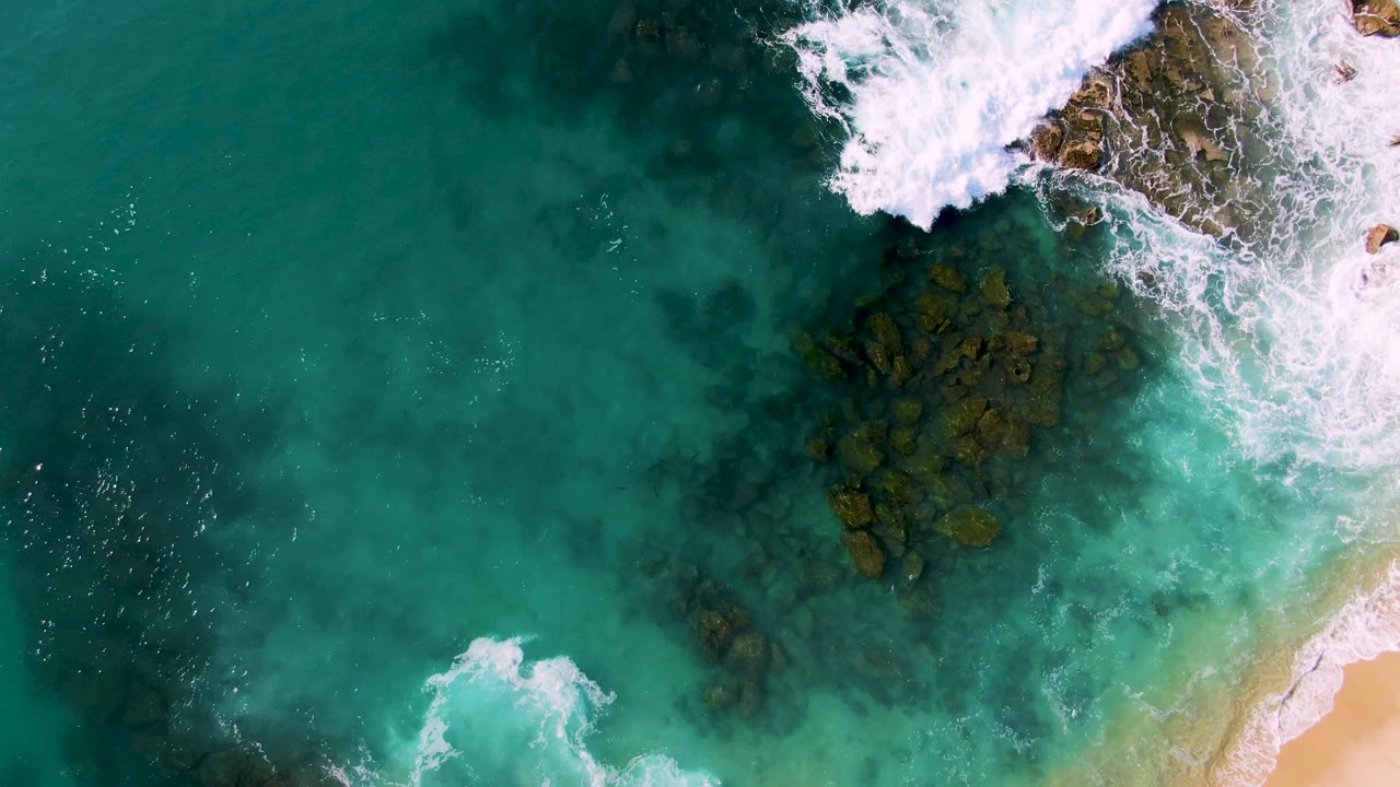 Aerial shoreline view with clear water, submerged rocks, white foam, and sand at the right edge.