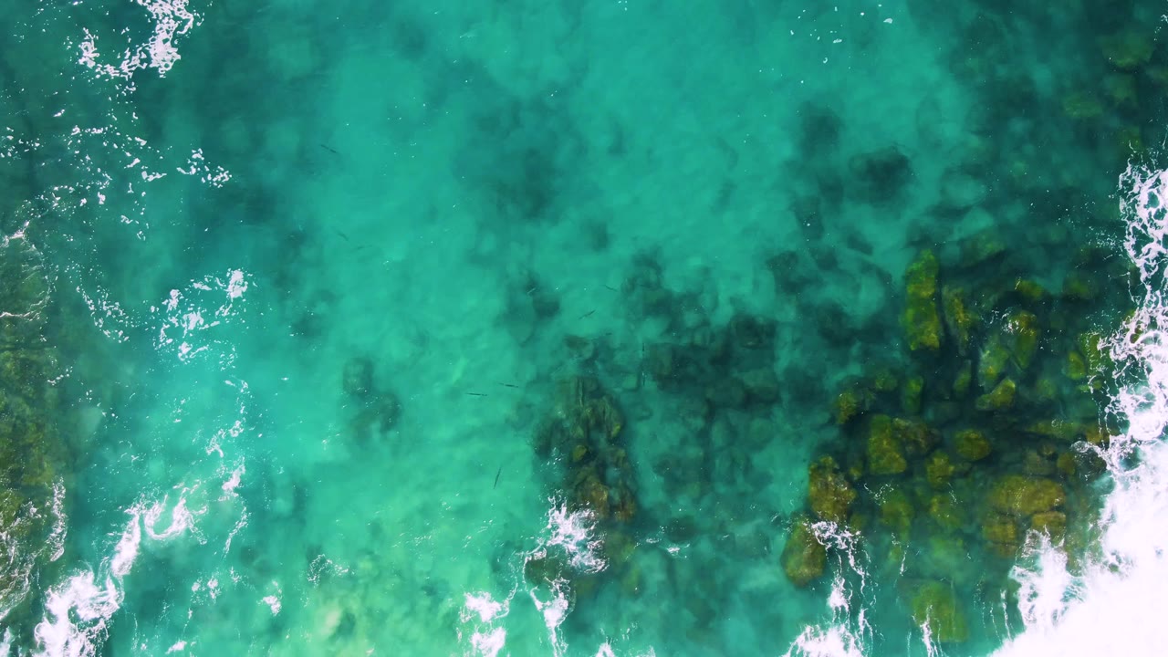 Aerial frame over Buzzards Beach East Cape BCS Mexico, where foamy white water crosses the lower center and dark rock patches sit beneath the turquoise surface, no visible people.