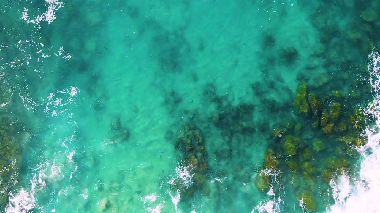 Top-down view at Buzzards Beach East Cape BCS Mexico, showing a larger dark rock cluster along the right side under clear turquoise water with white foam curling nearby, no visible people.