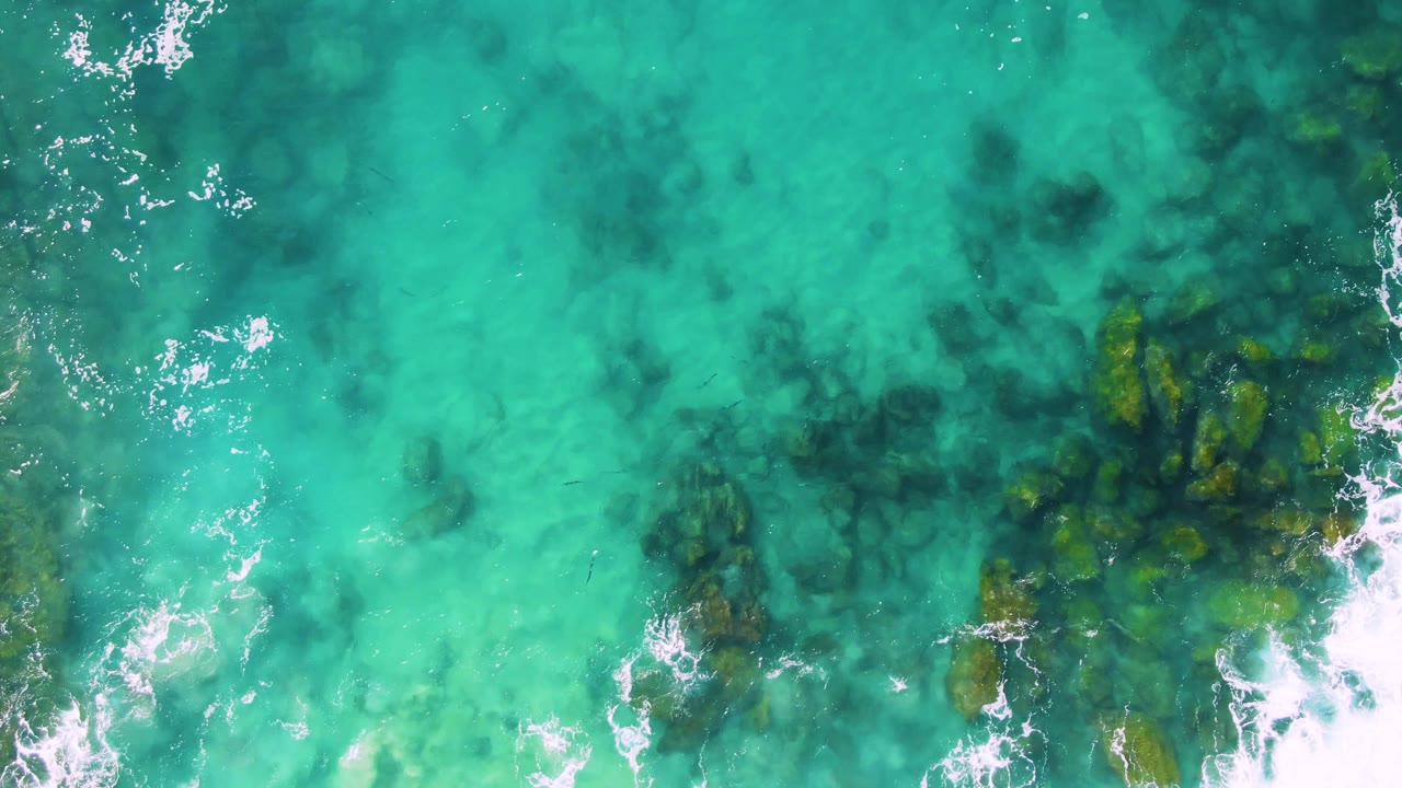 Aerial still from Buzzards Beach East Cape BCS Mexico with the submerged rock area lower in frame, pale turquoise shallows around it, and small white foam breaks, no visible people.