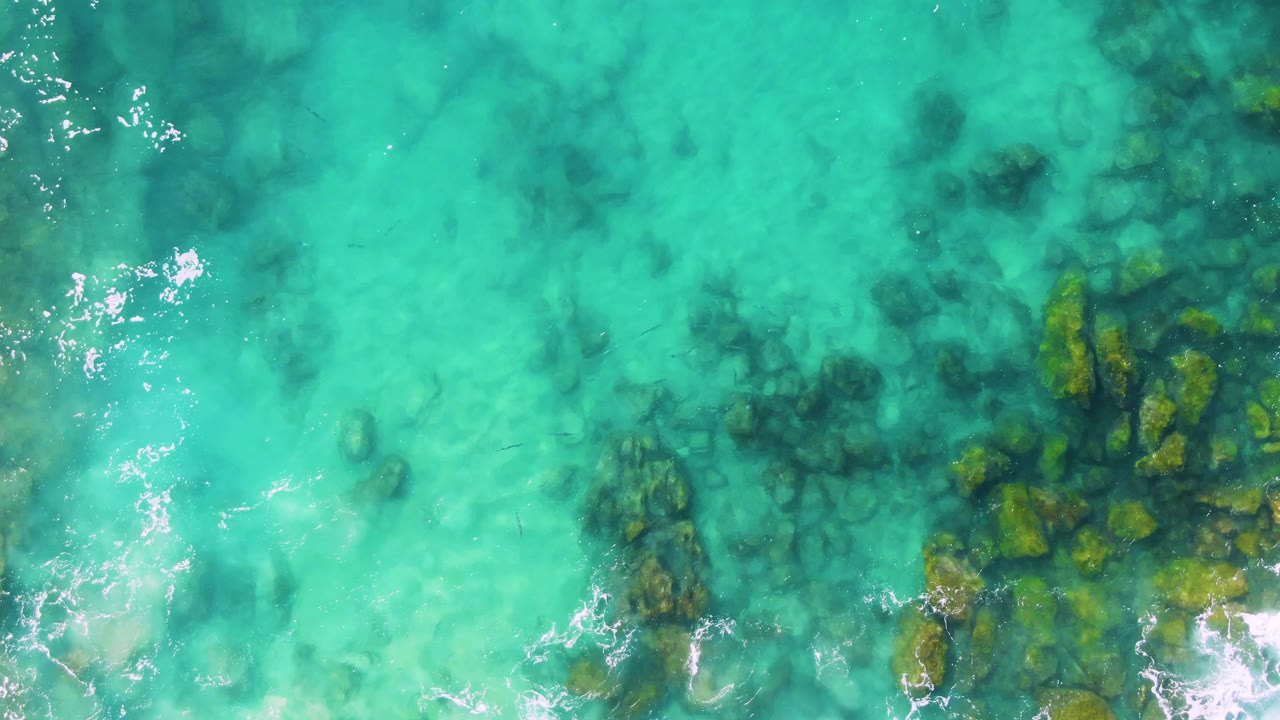 Top-down turquoise water over submerged rocks and small foam patches.