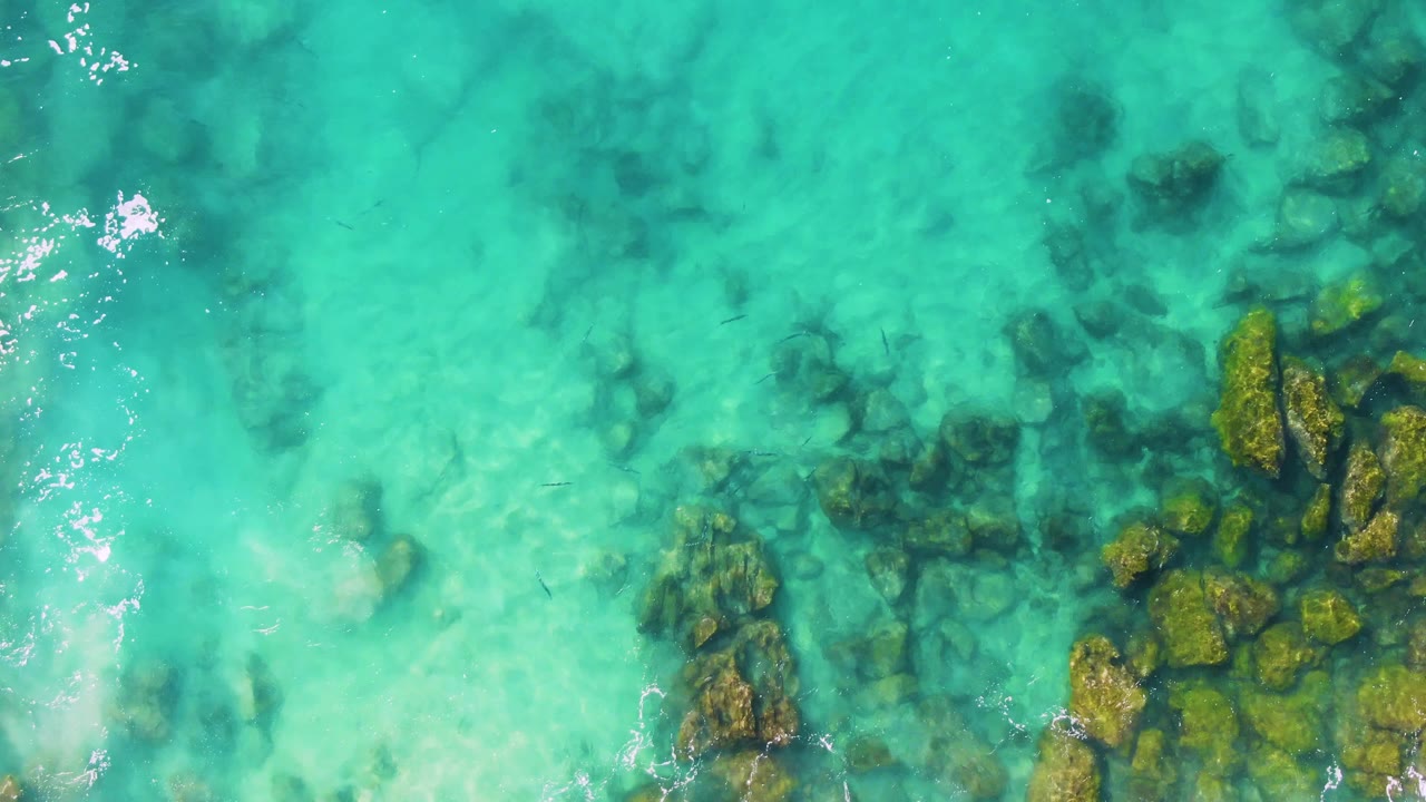 Overhead view at Buzzards Beach East Cape BCS Mexico with dark submerged rocks under blue-green water and a brighter foam line along the left side, with no visible people.