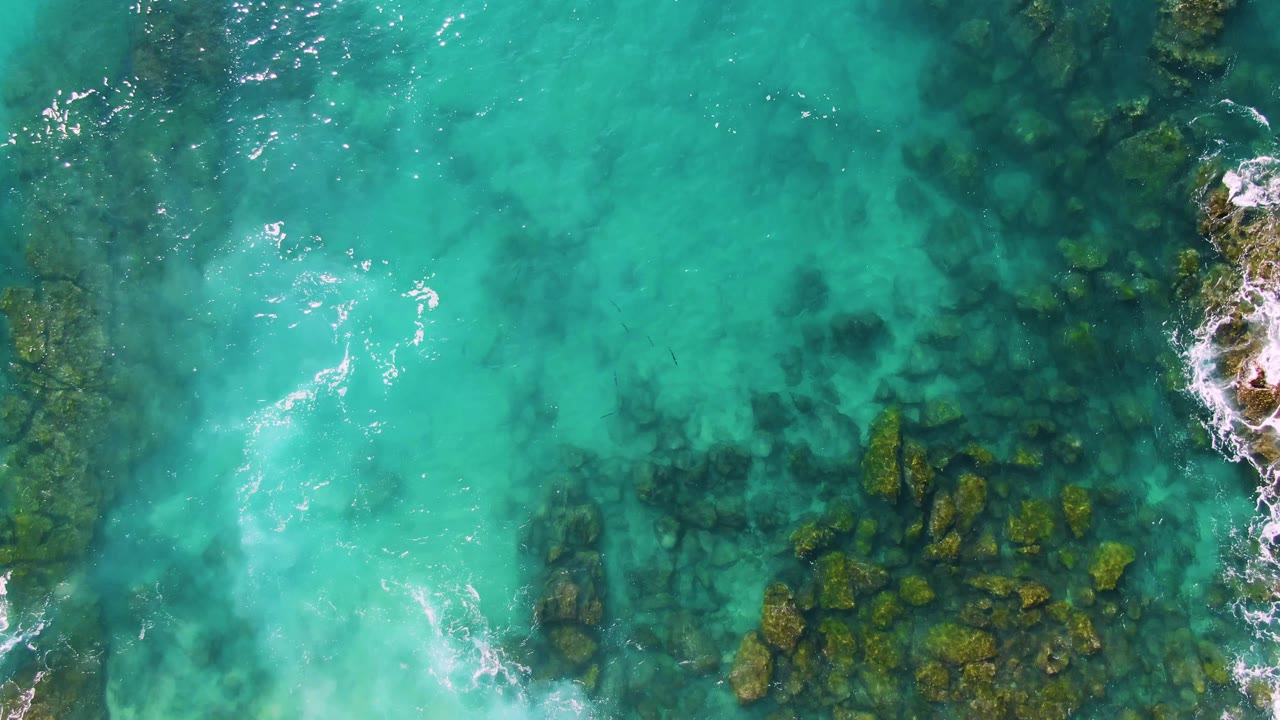 Second overhead frame at Buzzards Beach East Cape BCS Mexico with green algae-covered rocks clustered under clear turquoise water and small white ripples at the surface, with no visible people.