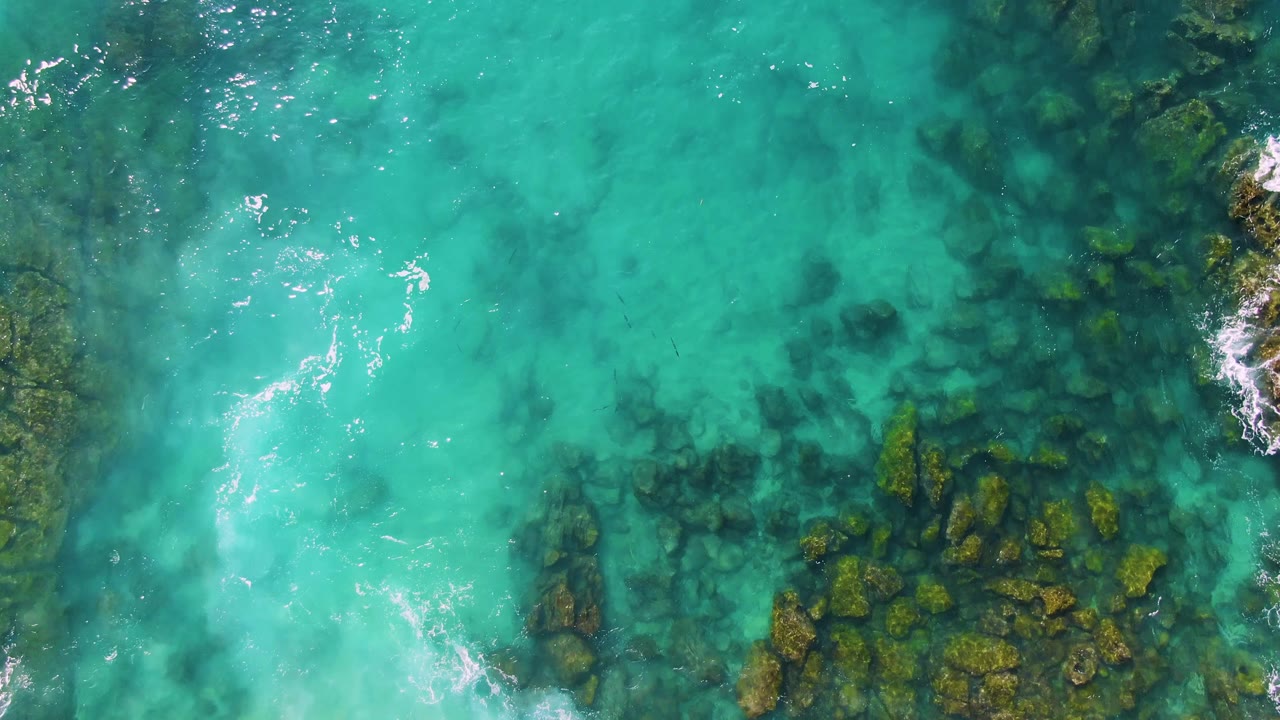 Overhead frame at Buzzards Beach East Cape BCS Mexico where white foam streaks cross the turquoise surface above darker rock patches, with no visible people.