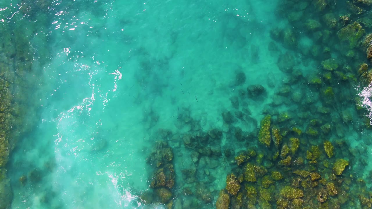 Mid-clip overhead view at Buzzards Beach East Cape BCS Mexico with blue-green shallow water, darker seabed shapes, a vertical line of submerged rocks, and no visible people.