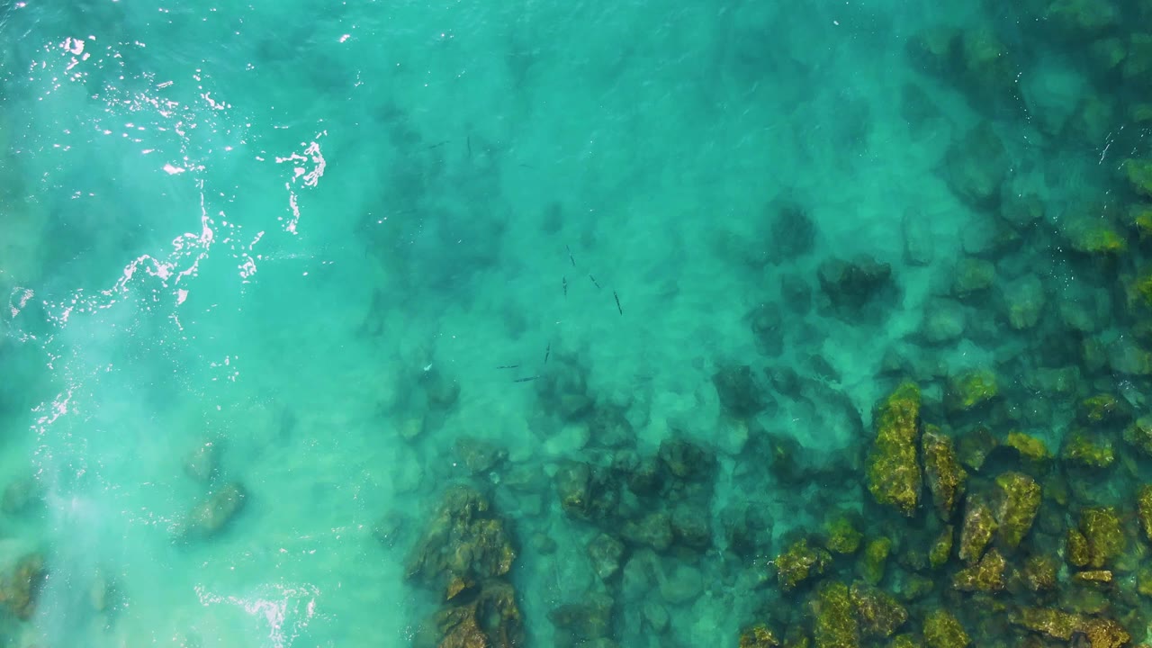 Top-down frame from Buzzards Beach East Cape BCS Mexico showing clear turquoise water, a rock cluster along the lower right, darker underwater patches, bright ripple highlights, and no visible people.
