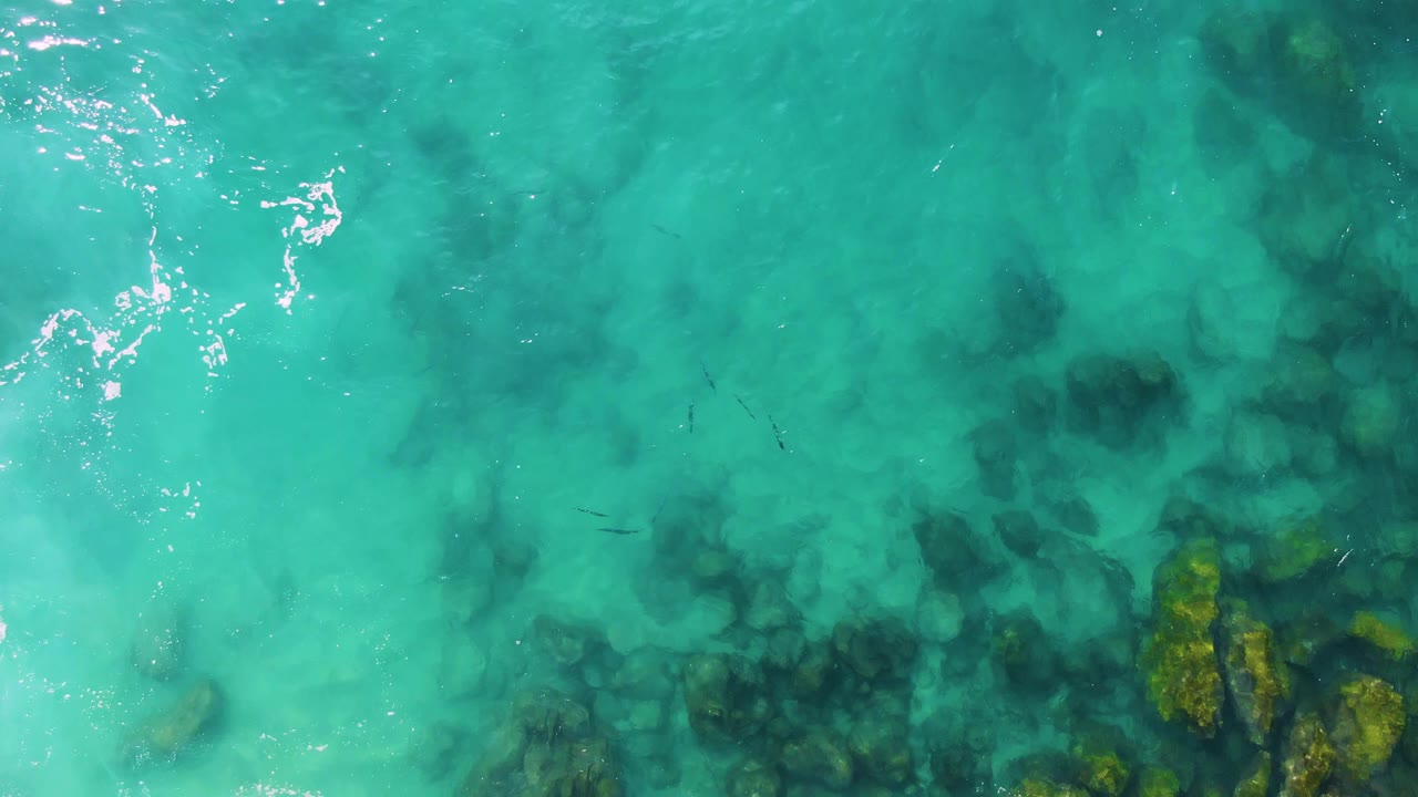 Buzzards Beach East Cape BCS Mexico seen from above, with turquoise water, darker submerged patches through the center, rocks along the lower right, light surface ripples, and no visible people.