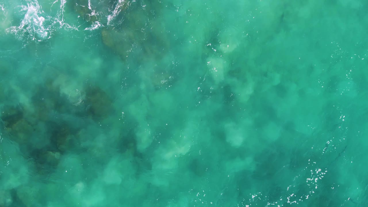 Overhead view of turquoise water at Buzzards Beach East Cape BCS Mexico, with white foam near the upper edge and dark submerged rocks below, no visible people.