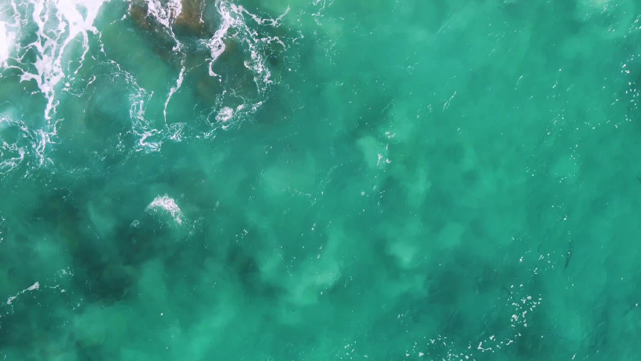 Top-down view at Buzzards Beach East Cape BCS Mexico where white foam breaks across the upper frame over turquoise water and submerged rock shadows, no visible people.