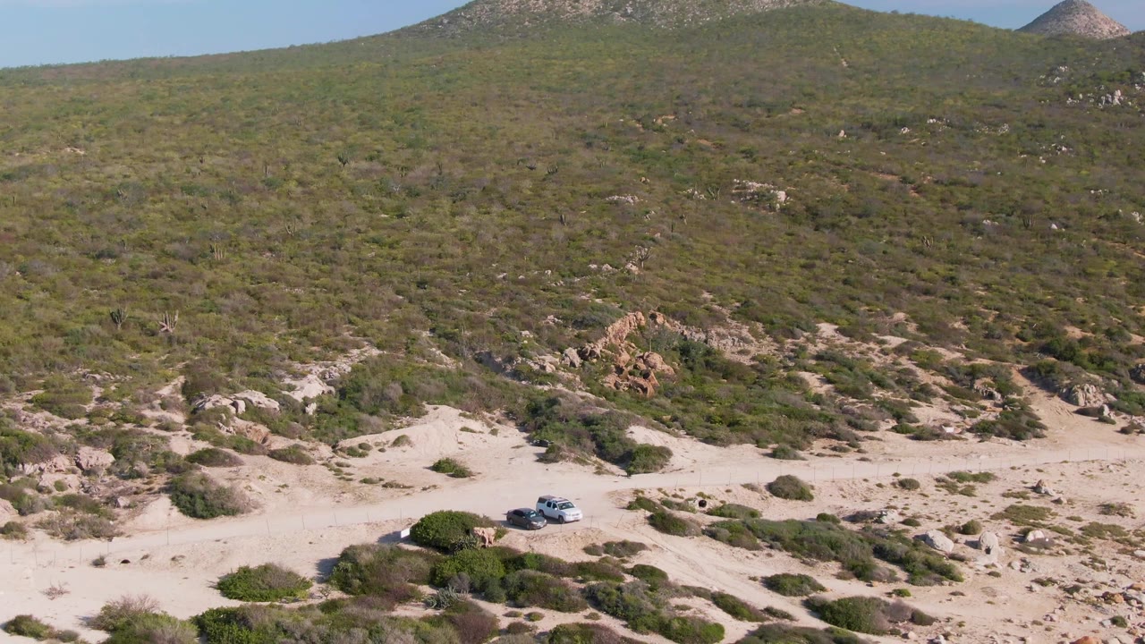 A white SUV sits low in the frame on a sandy off-road trail in Buzzards Beach East Cape BCS Mexico, with green shrubs and rocky hillside behind it and no visible people.