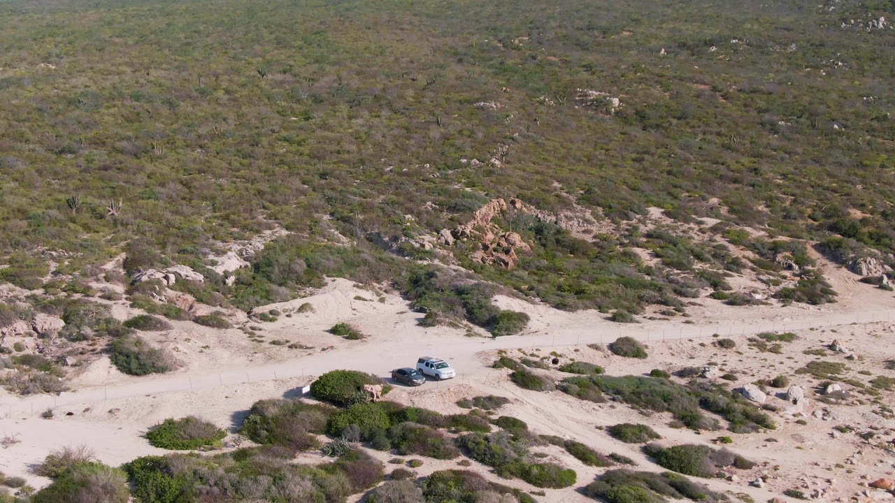 The white SUV appears farther along the pale track in Buzzards Beach East Cape BCS Mexico, surrounded by low shrubs, beige sand, and rocky scrub terrain with no visible people.