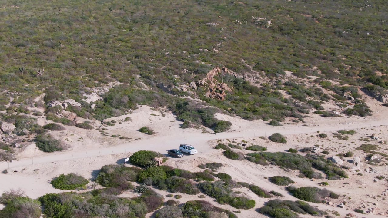 A drone-height view shows the white SUV on the sandy road in Buzzards Beach East Cape BCS Mexico, with clustered shrubs, exposed rocks, and a scrubby hillside behind it and no visible people.